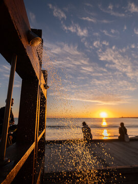 View of Bathers Beach with a wooden outdoor shower spraying water at sunset, with people sitting on the deck overlooking the ocean in City of Fremantle, Western Australia, Australia.