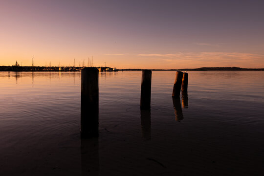 View of wooden posts in the calm water at sunset with a distant marina and soft orange sky at Emu Point, Western Australia, Australia.