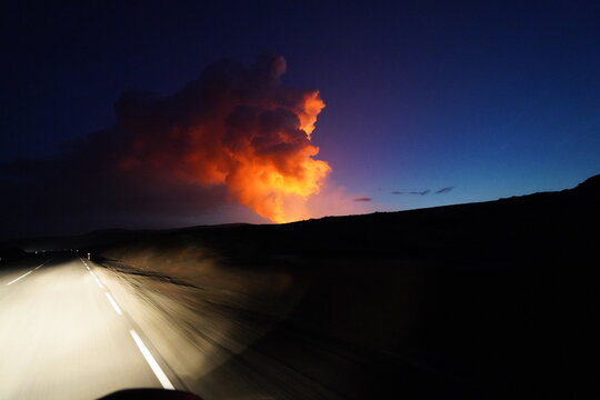 View of Fagradalsfjall Volcano erupting at night with glowing lava plumes rising into the dark sky above a road in Grindavik, Grindavikurbaer, Iceland.