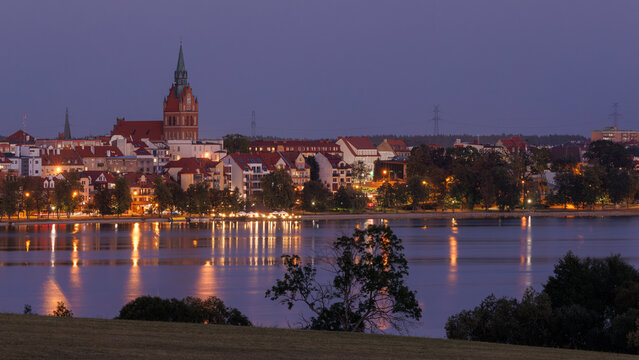 View of the illuminated waterfront and church spire reflecting on the calm lake at twilight in Elk, Warmian-Masurian Voivodeship, Poland.