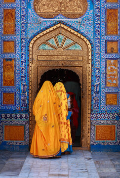 View of the Shrine of Sachal Sarmast with women in yellow traditional clothing entering the ornate blue and gold tiled entrance in Daraza Sharif, Sindh, Pakistan.