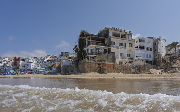 View of beachfront buildings with balconies and stone foundations overlooking the sandy beach and ocean waves in Taghazout, Souss-Massa, Morocco.