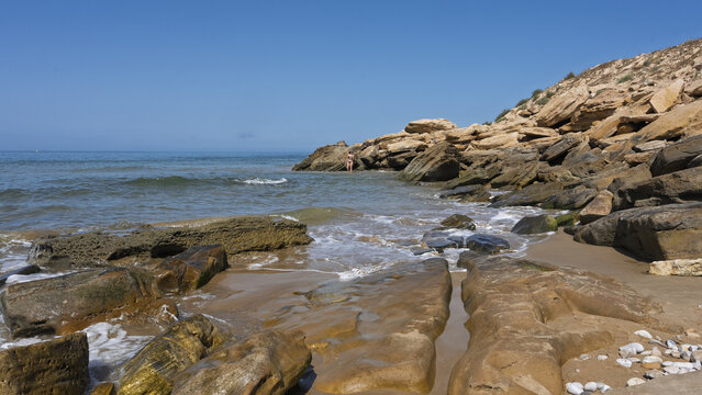 View of a rocky coastline with gentle waves washing over sandstone formations and a person standing near the water under a clear blue sky in Taghazout, Souss-Massa, Morocco.