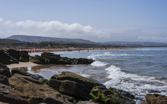 View of the sandy beach with rows of red and white umbrellas, rocky shoreline in the foreground, and calm ocean waves Taghazout, Souss-Massa, Morocco.