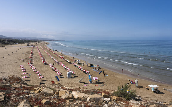 View of a sandy beach lined with rows of red and white umbrellas, surfboards, and people enjoying the sunny weather along the Atlantic coastline in Taghazout, Souss-Massa, Morocco.