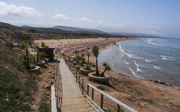 View of the sandy beach with rows of parasols, palm trees along the wooden walkway, and gentle waves rolling onto the shore in Taghazout, Souss-Massa, Morocco.