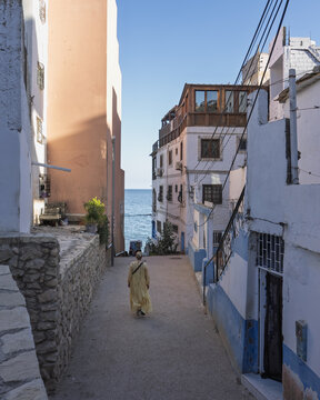 View of a person in a yellow dress walking down a narrow street between traditional white and peach-colored buildings towards the blue ocean in Taghazout, Souss-Massa, Morocco.