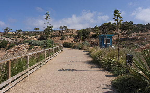 View of a paved coastal path lined with a wooden fence and agave plants, featuring a small blue guard shack in a dry landscape under a clear blue sky in Taghazout, Souss-Massa, Morocco.
