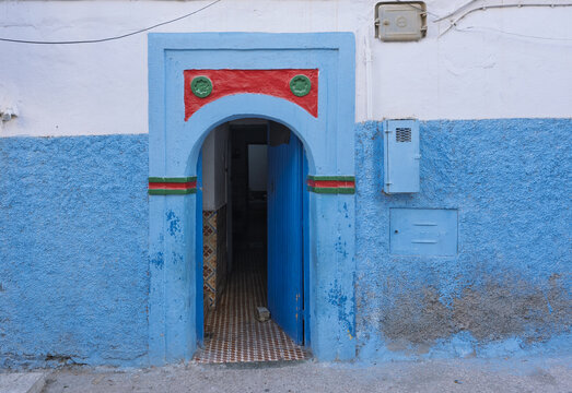 View of a traditional blue-painted doorway with a red and green arched frame set into a textured wall in Taghazout, Souss-Massa, Morocco.