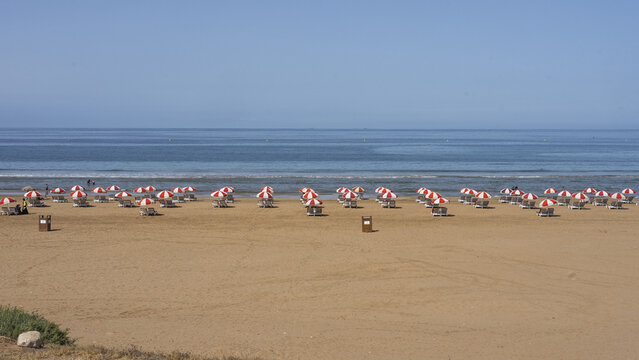 View of red and white striped beach umbrellas lined up on the sandy shore overlooking the Atlantic Ocean in Taghazout, Souss-Massa, Morocco.