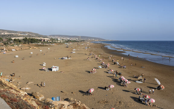 View of the sandy beach with colorful umbrellas and people relaxing along the coastline in Taghazout, Souss-Massa, Morocco.