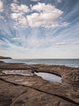View of the rocky coastline and tide pools at Royal National Park under a vast sky with wispy clouds in Royal National Park, New South Wales, Australia.
