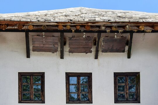 View of a traditional house facade with a stone roof, wooden support beams, and three dark-framed windows against a white plastered wall in Bosnia and Herzegovina.