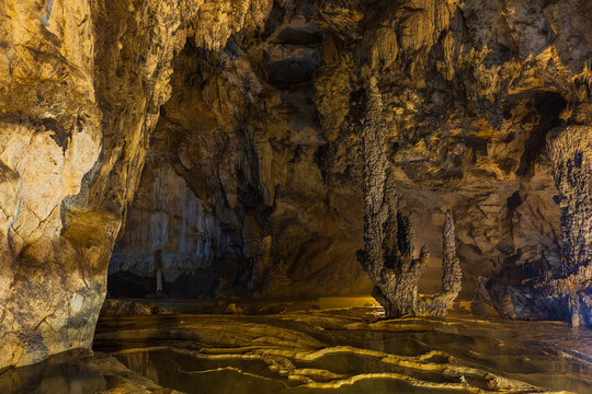 View of Nguom Ngao Cave with limestone stalactites and stalagmites, illuminated by soft lighting, reflecting in calm water pools on the cave floor in Dam Thuy, Cao Bang, Vietnam.