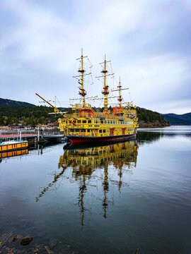 View of the yellow sightseeing pirate ship on Lake Ashinoko with its reflection in the calm water, surrounded by forested mountains in Hakone, Kanagawa, Japan.