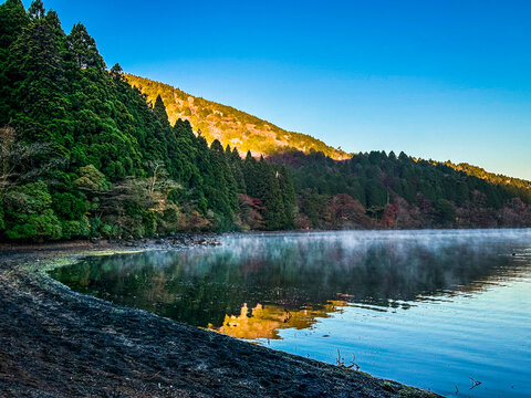 View of Lake Ashinoko with mist rising from the water, surrounded by dense evergreen forests on the hills under a clear blue sky in Hakone, Kanagawa, Japan.