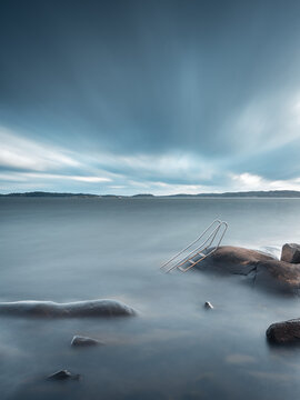 View of a metal swimming ladder attached to a rock on the calm coastline of Fiskeback under a dramatic cloudy sky in Gothenburg, Vastra Gotaland County, Sweden.