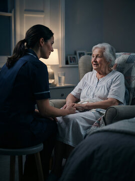 Night nurse holding an elderly woman's hands beside her armchair