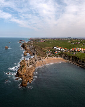 Aerial view of Arnia Beach with its dramatic rock formations, sandy shoreline, and green coastal fields overlooking the Cantabrian Sea in Cantabria, Spain.