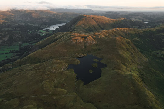 Aerial view of Angletarn Pikes with Angle Tarn, rolling hills, and distant lake under soft golden hour light in Penrith, England, United Kingdom.