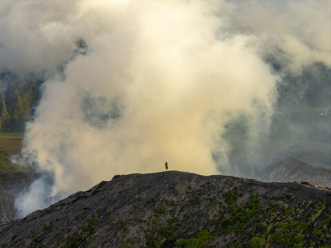 Aerial view of a lone person standing on the rugged rim of Mount Bromo with thick volcanic smoke billowing into the sky in Tosari, East Java, Indonesia.