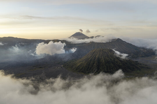 Aerial view of Mount Bromo with its volcanic crater emitting smoke, surrounded by clouds and rugged mountain landscape in Tosari, East Java, Indonesia.