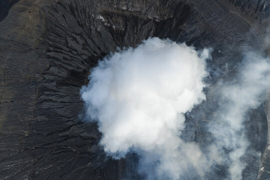 Aerial view of Mount Bromo crater with thick volcanic smoke rising from the rugged dark terrain in Tosari, East Java, Indonesia.