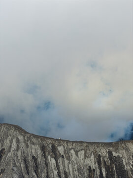 Aerial view of the rugged crater rim of Mount Bromo with volcanic smoke rising into the sky and people walking along the ridge in Tosari, East Java, Indonesia.