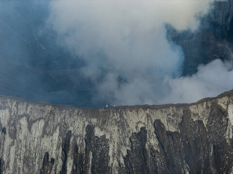 Aerial view of the Mount Bromo crater rim with people walking along the edge, surrounded by volcanic smoke and rugged terrain in Tosari, East Java, Indonesia.