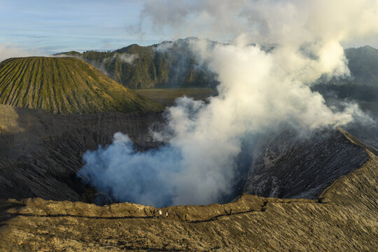 Aerial view of Mount Bromo crater with thick volcanic smoke rising from the rugged caldera and people walking along the rim in Tosari, East Java, Indonesia.