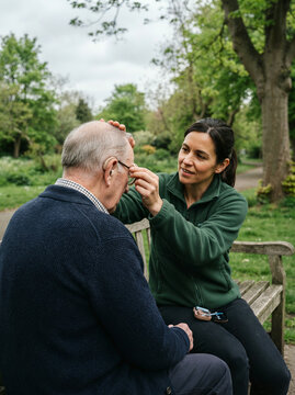 Younger woman adjusting the hearing aid of an older man on a park bench