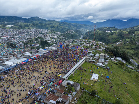 Aerial view of the Giant Kites of Sumpango festival with colorful kites, crowds of people in the cemetery, surrounding hills, and cloudy sky in Sumpango, Sacatepequez, Guatemala.