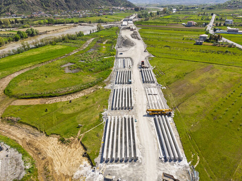 Aerial view of a construction site with rows of concrete pillars for a new bridge project, a yellow crane, and rural fields in Paper, Elbasan County, Albania.