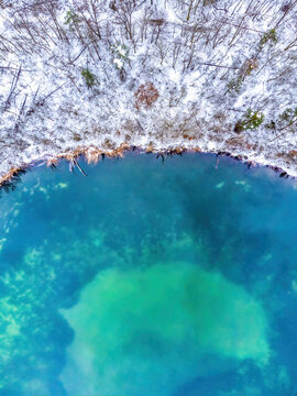 Aerial view of a turquoise lake bordered by a snow-covered forest with bare trees in winter in Mannheim, Baden-Wurttemberg, Germany.