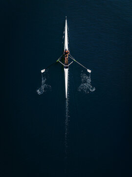 Aerial view of a lone rower in a scull boat gliding across dark water, creating ripples with oars in Mannheim, Baden-Wurttemberg, Germany.