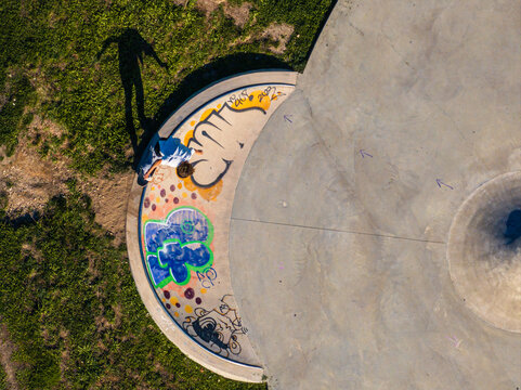 Aerial view of a skateboarder performing tricks in a concrete skatepark with colorful graffiti on the curved ramp and a long shadow cast on the grass in Mannheim, Baden-Wurttemberg, Germany.
