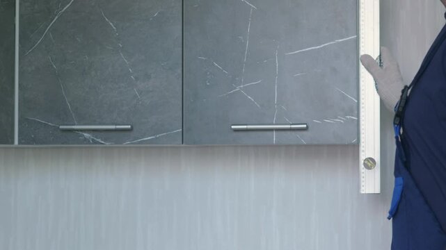 A handyman neatly closes the door of a modern dark kitchen cabinet during installation. Afterwards, the professional worker uses a spirit level to check the vertical alignment 
