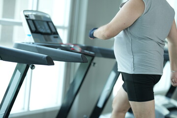 Fototapeta premium Overweight middle aged man in gray cap and sleeveless shirt walks the treadmill in close up, step after heavy step at the start of his new healthy lifestyle in the gym in warm natural light