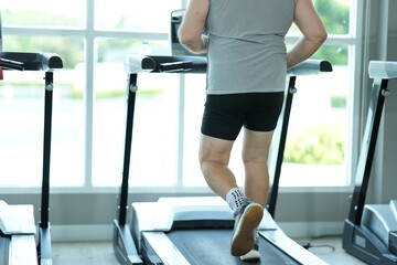 Fototapeta premium Overweight middle aged man in gray cap and sleeveless shirt puts one foot before the other on the moving belt during his weight loss workout at the fitness club, candid real moment