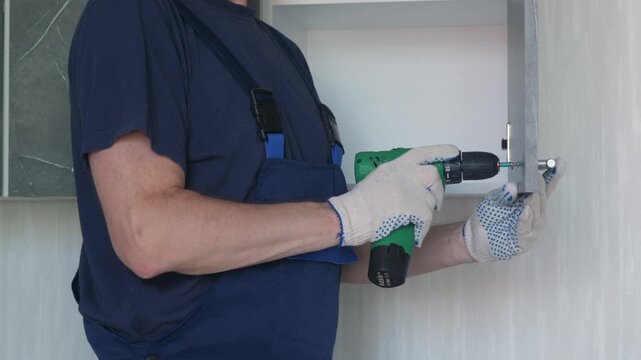 A handyman in a blue uniform and protective gloves uses a cordless screwdriver to attach a handle to a kitchen cabinet door. 