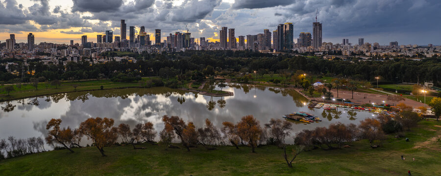 Aerial view of Yarkon Park with its lake and lush greenery, featuring the modern Tel Aviv skyline under a dramatic sunset sky in Tel Aviv-Yafo, Tel Aviv District, Israel.