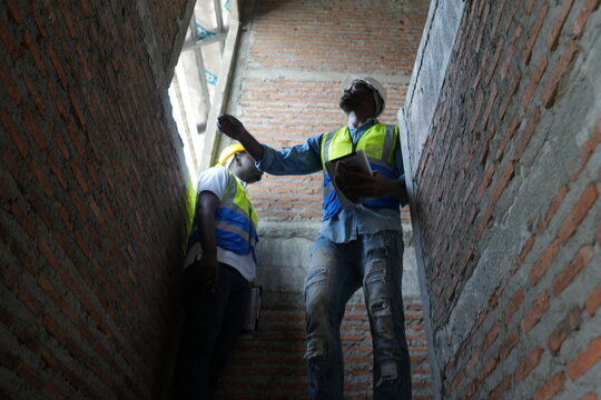 African foreman and young worker in safety vests test the strength of the masonry with their hands inside the bare brick shell of a new home under construction with copy space