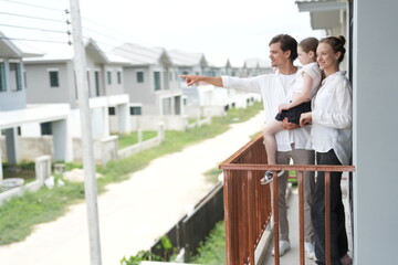 Obraz premium Young parents holding their child look out over the rows of new white houses from the railing on the upper balcony of their new home above the neighborhood in warm natural light