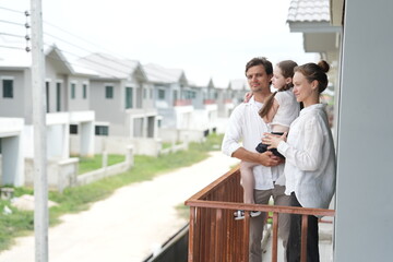 Obraz premium Young parents holding their child stand at the wooden railing admiring the neighborhood view on the upper balcony of their new home above the neighborhood on a bright sunny day