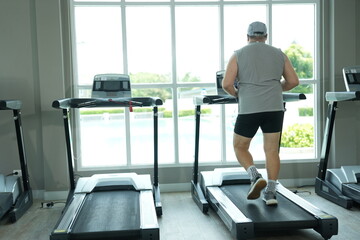 Fototapeta premium Overweight middle aged man in gray cap and sleeveless shirt raises his arms in victory while standing on the treadmill in a bright modern gym with large windows