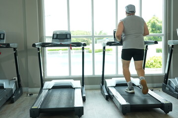 Fototapeta premium Overweight middle aged man in gray cap and sleeveless shirt raises his arms in victory while standing on the treadmill at the start of his new healthy lifestyle in the gym
