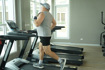 Fototapeta premium Heavyset mature man determined to lose weight does his cardio walk on the treadmill machine in a bright modern gym with large windows. Funny weight loss and healthy lifestyle concept