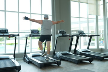 Fototapeta premium Funny fat senior man in sportswear balances playfully on the treadmill with arms spread wide during his weight loss workout at the fitness club in warm natural light