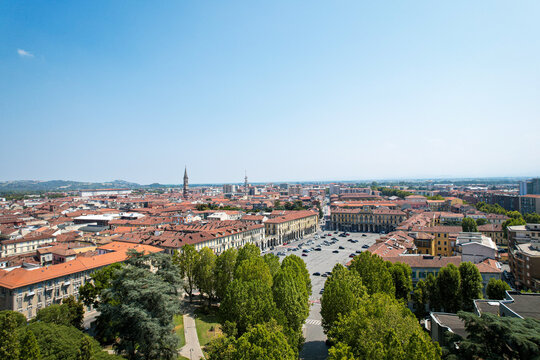 Aerial view of Piazza della Liberta featuring historic buildings, a large open square with parked cars, and surrounding trees under a clear blue sky in Alessandria, Piedmont, Italy.