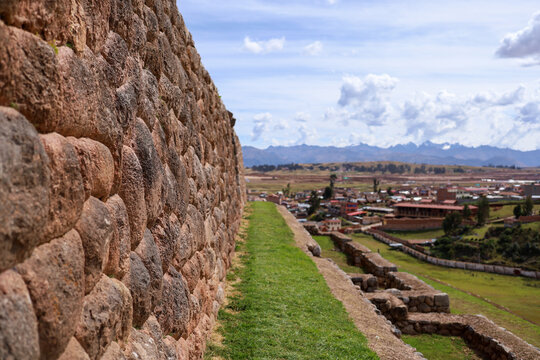 View of the ancient Inca stone wall at Chinchero with a grassy path and a view of the town and mountains in the background in Cusco, Cusco, Peru.